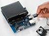 Close-up of the Arduino VENTUNO Q board with heatsink, showing a hand plugging a USB cable into the port beside a camera module.
