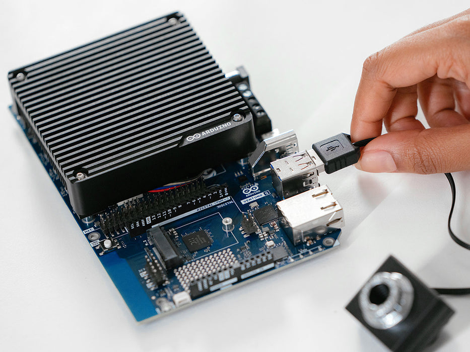 Close-up of the Arduino VENTUNO Q board with heatsink, showing a hand plugging a USB cable into the port beside a camera module.