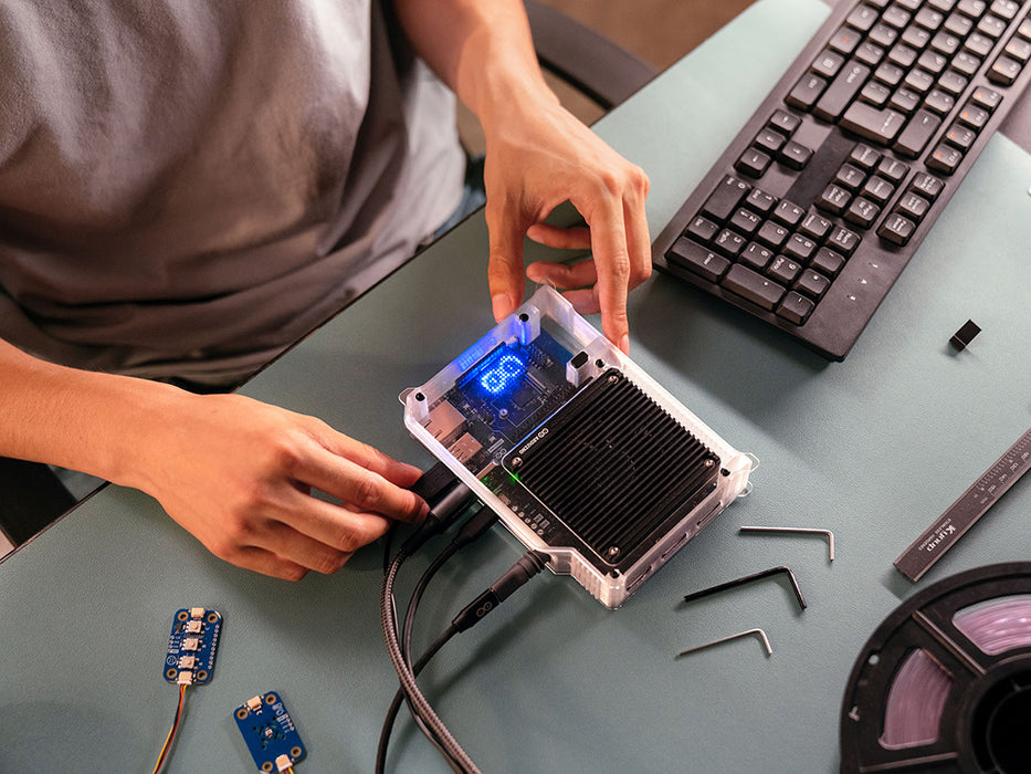 Close-up of hands assembling the Arduino VENTUNO Q into a clear case on a desk, with a lit blue display and Modulino boards nearby.