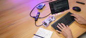 High-angle lifestyle photograph of an organized electronics workstation showcasing a completed DIY project from the Arduino UNO Q Arcade Bundle. A person's hands are typing on a grey keyboard. On the light wood desk, a custom, 3D-printed handheld game controller enclosure—integrating the UNO Q board, joystick, and buttons—is connected via USB-C to an Arduino-branded USB-C hub and a small external monitor displaying project code. A notebook with notes, a pencil, and a mouse complete the setup.