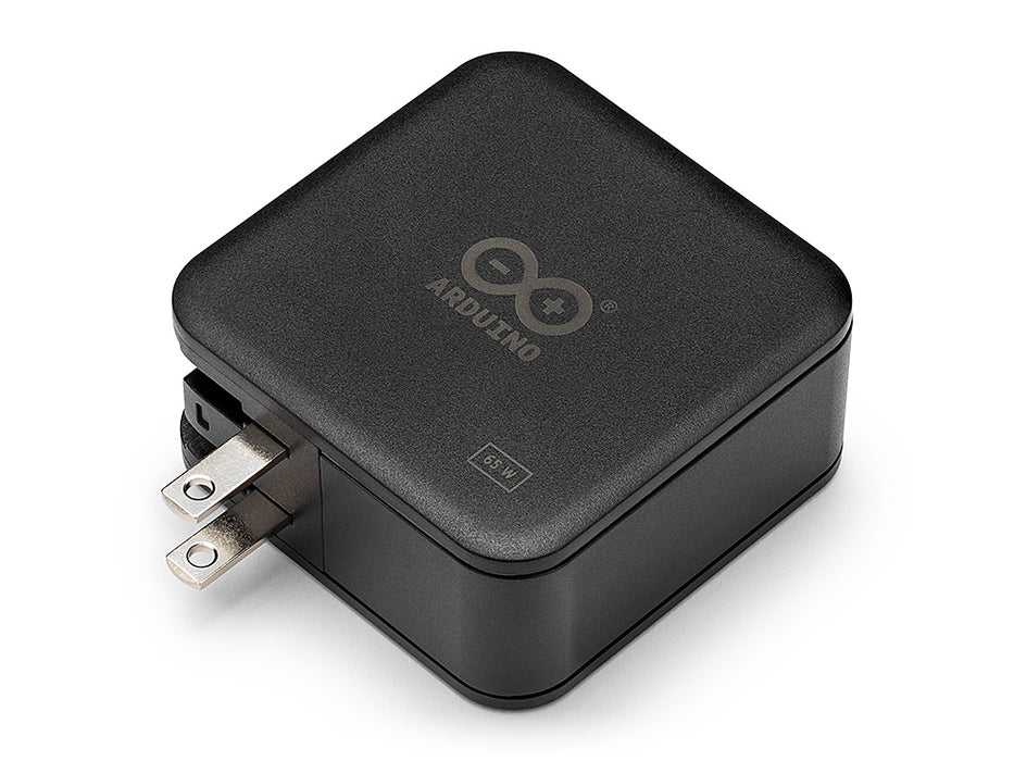 Angled top view of the black Arduino 65W power adapter showing the grey logo and the extendable US-style power plug on the side.